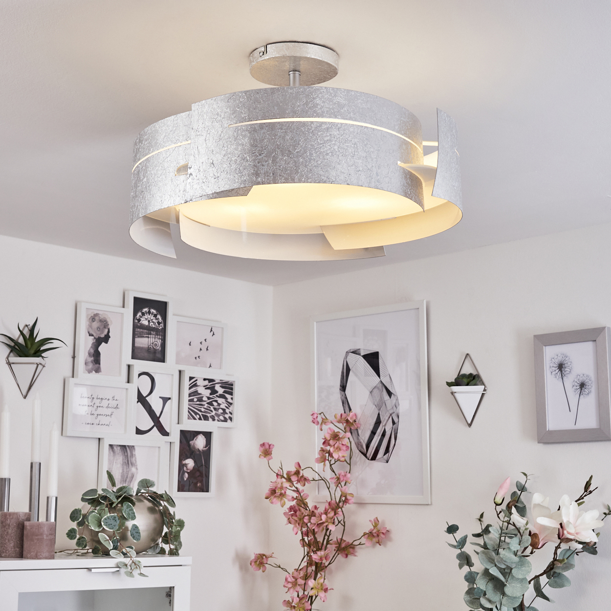 Eine silberne Deckenleuchte beleuchtet einen weißen Raum. Die Einrichtung umfasst gerahmte Kunst, Pflanzen und Blumen, was ein modernes Interieur schafft.