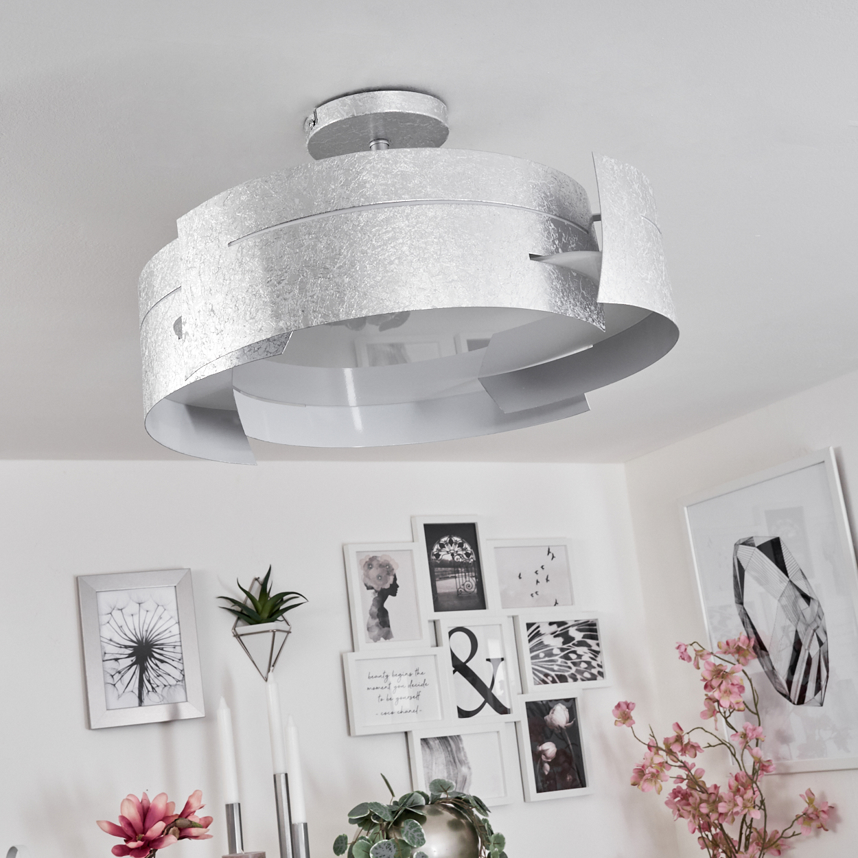 Silberne Deckenlampe mit strukturierter Oberfläche. Darunter weiße Wände mit gerahmter Kunst und Blumen. Ein modernes, helles Interieur.