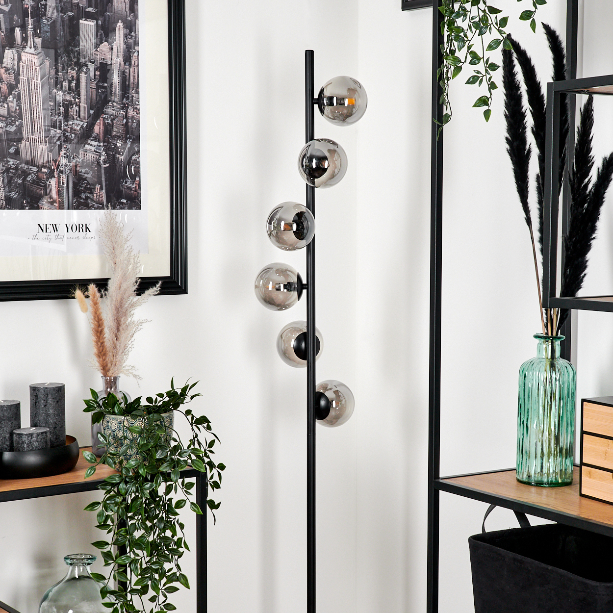 Ein moderner Raum mit einer schwarzen Lampe mit runden Glühbirnen, einem New York-Druck, Pflanzen, Regalen und Kerzen.