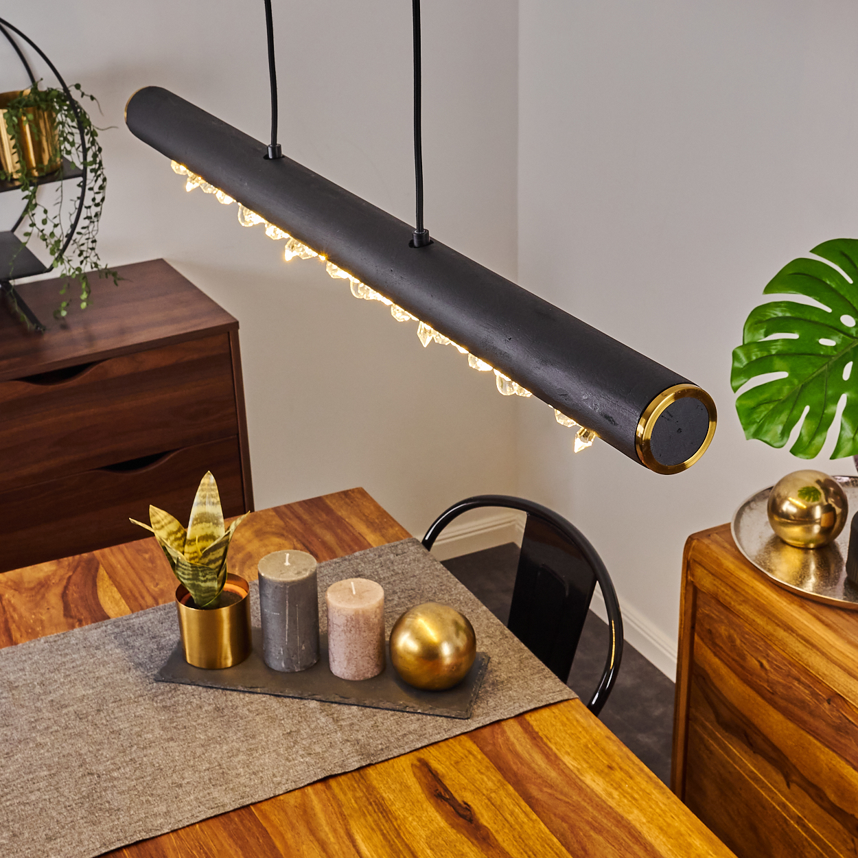 Schwarze, goldverkleidete Pendelleuchte über einem Holztisch. Kerzen, Pflanze und eine goldene Kugel stehen auf dem Tisch. Holzmöbel im Hintergrund.