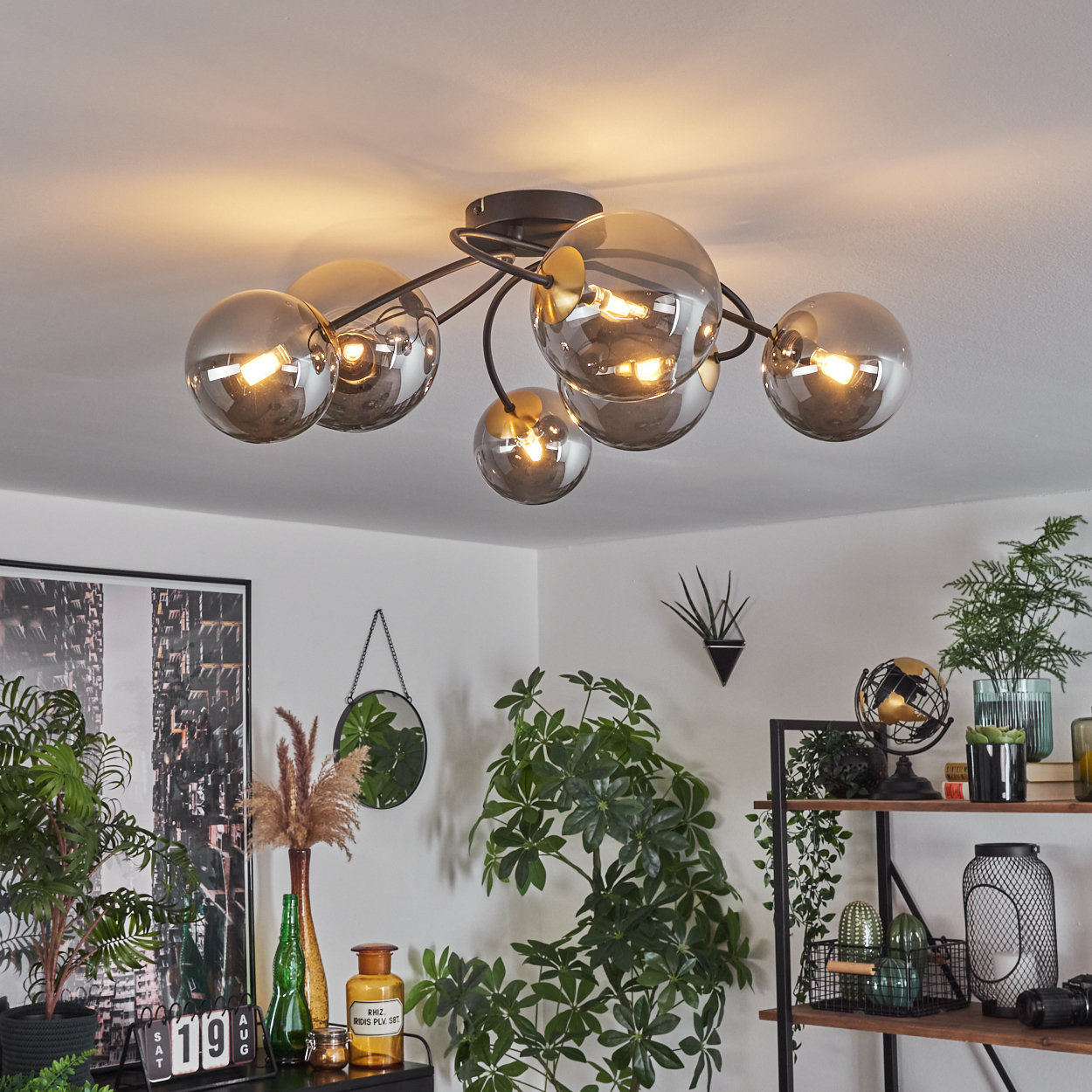 Eine Deckenleuchte mit Kugelschirmen beleuchtet den Raum. Pflanzen und Dekor sind sichtbar. Ein schwarz gerahmtes Bild hängt an der Wand.