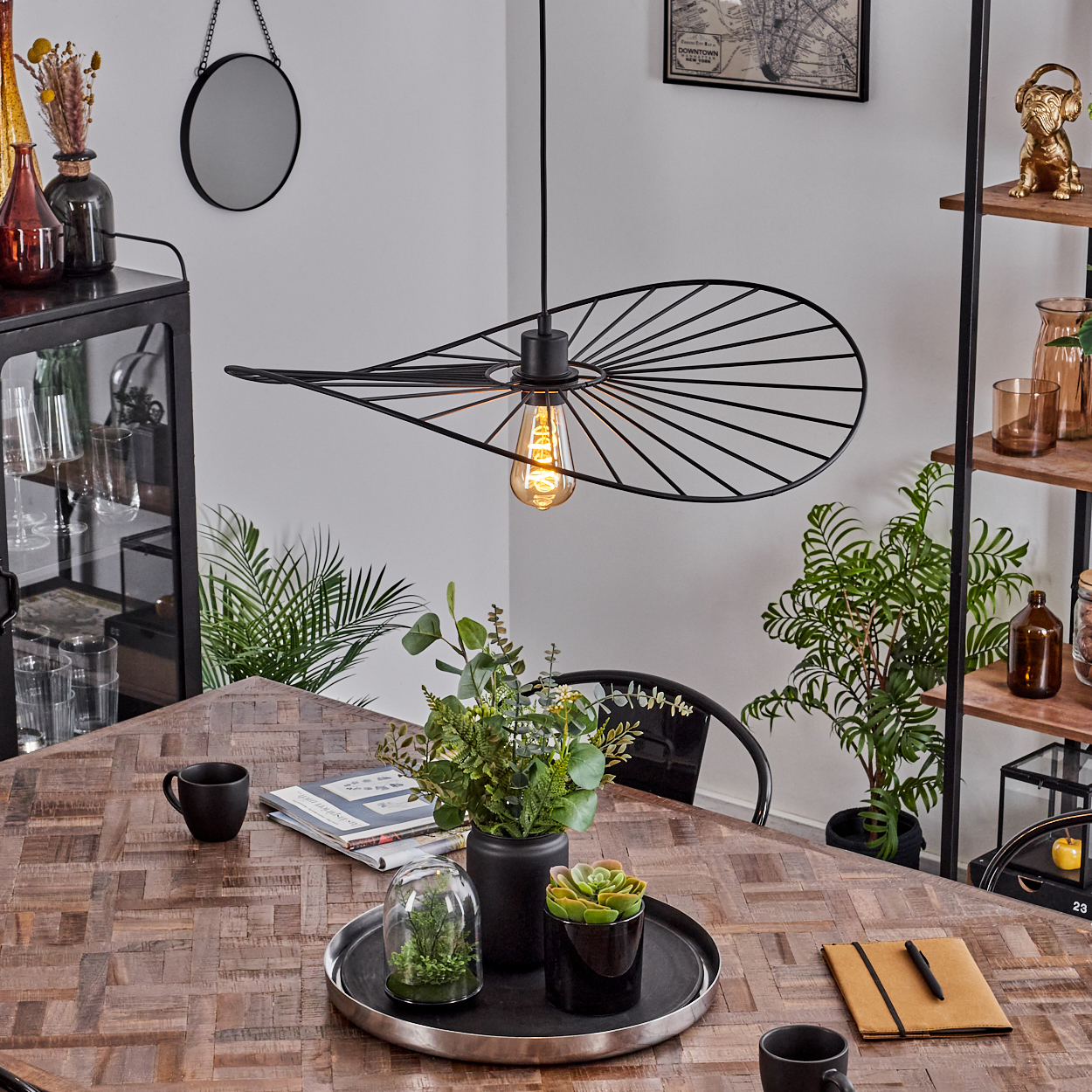 Ein modernes Esszimmer mit Holztisch, schwarzer Lampe, Pflanzen und Bücherregal. Auf dem Tisch ein Tablett mit Pflanzen.
