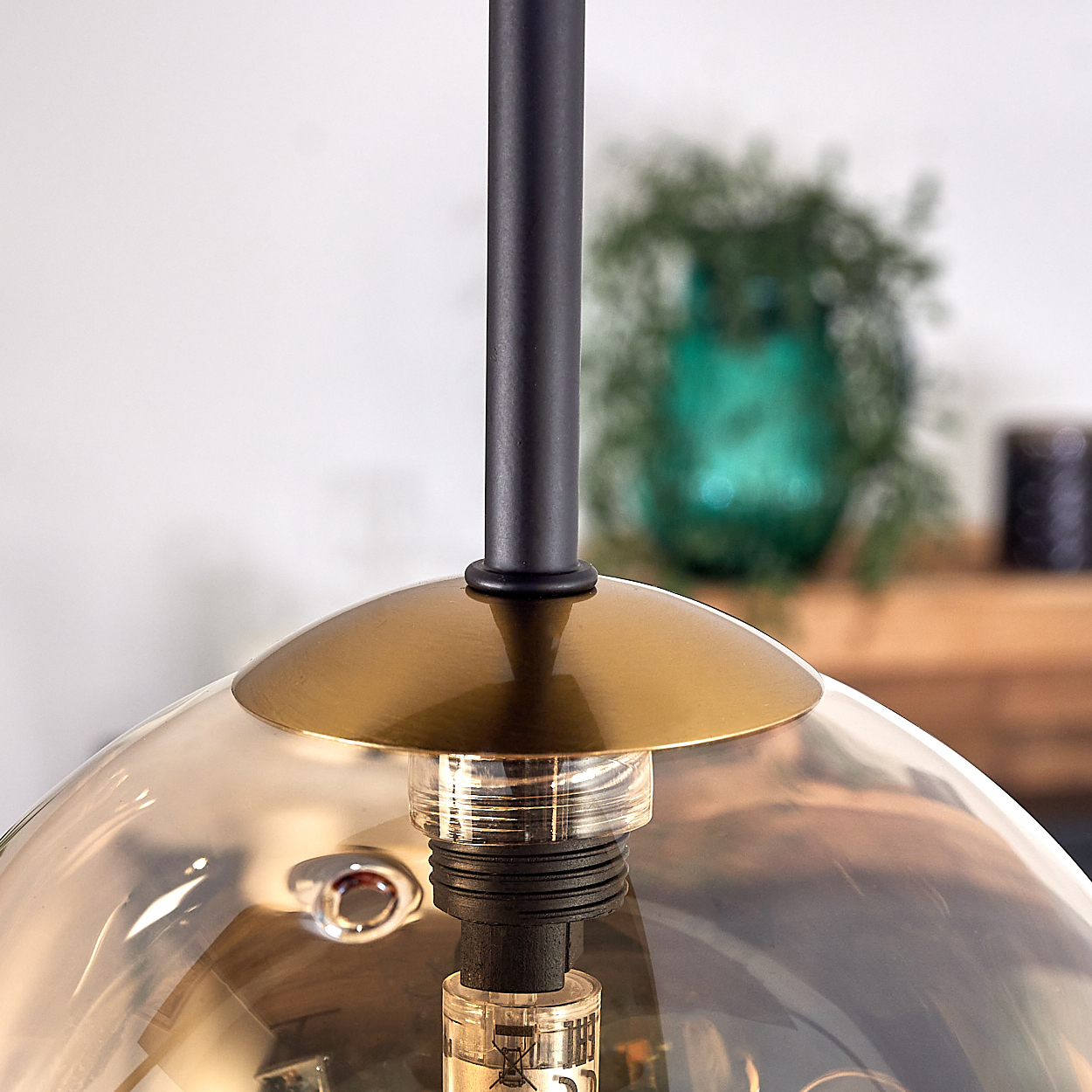 Nahaufnahme einer modernen Lampe. Sie hat eine schwarze Stange, einen goldfarbenen Schirm und eine transparente Glühbirne. Im Hintergrund ist eine grüne Pflanze.
