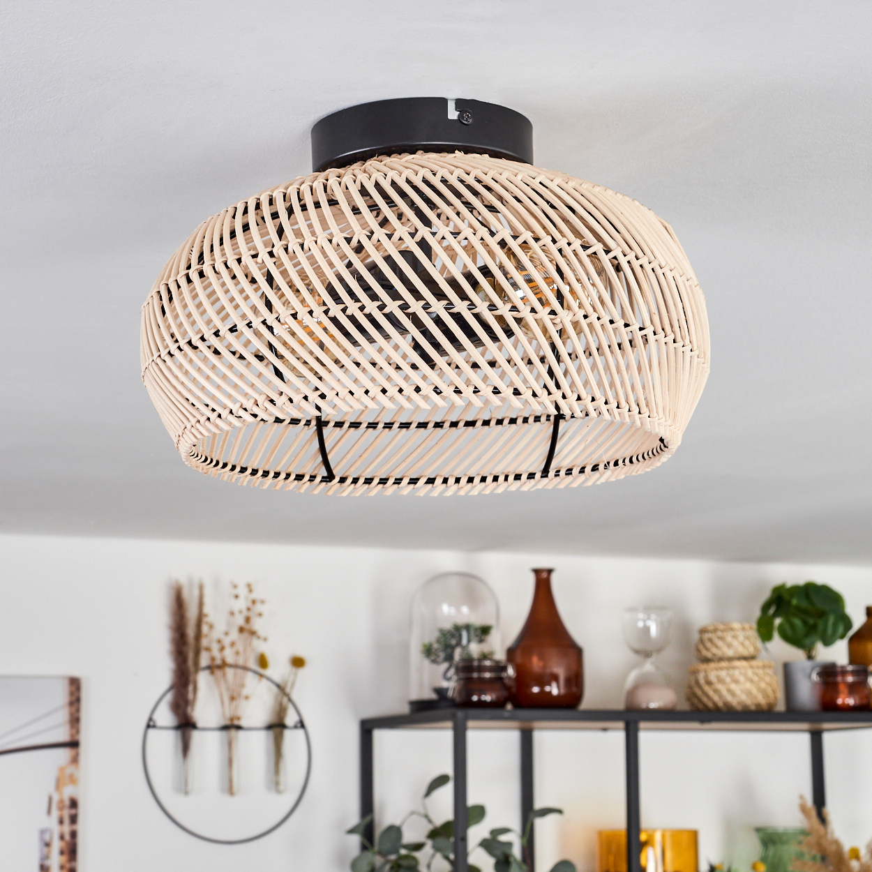 Geflochtene Deckenlampe aus Rattan. Schwarze Halterung. Regal mit Dekoration im Hintergrund: Vasen, Pflanzen und andere Dekoartikel.