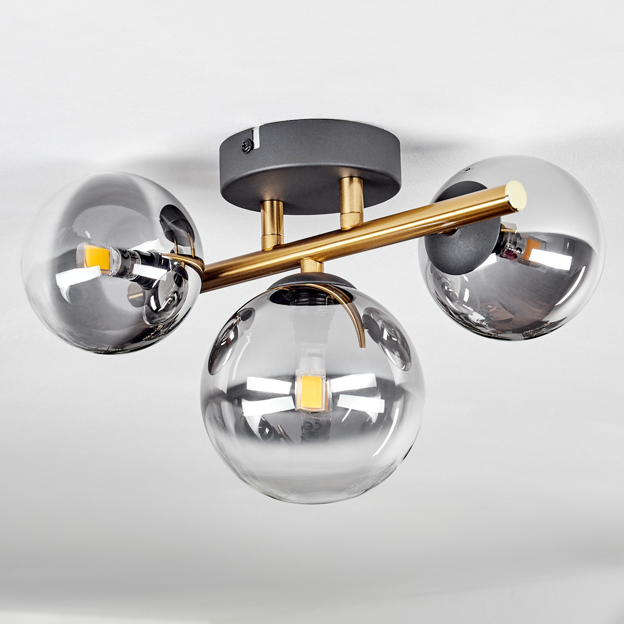 Eine moderne Deckenlampe mit drei kugelförmigen Glasschirmen, goldenen Armen und einem grauen Sockel. Die Glühbirnen sind sichtbar.