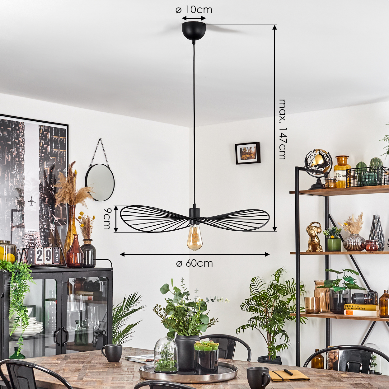 Eine schwarze Hängelampe hängt über einem Holztisch mit Pflanzen und Dekoration. Regale befinden sich im Hintergrund.