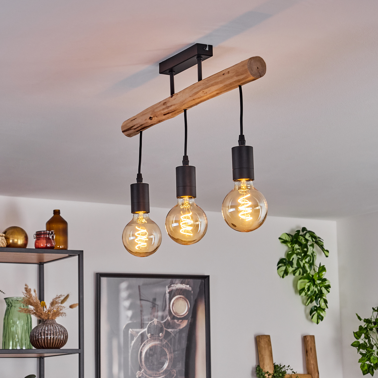 Ein Holzbalken mit drei hängenden Glühbirnen beleuchtet einen Raum. Ein Regal mit Dekorationen und ein gerahmtes Bild sind im Hintergrund.