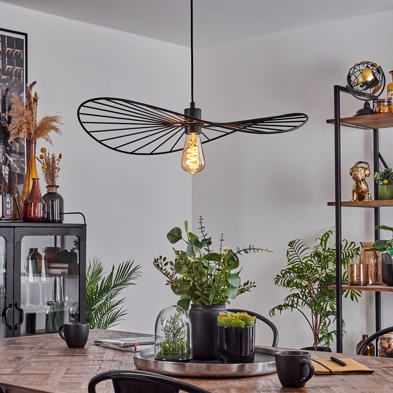 Ein Esszimmer mit Holztisch, schwarzen Stühlen und dekorativen Pflanzen. Eine schwarze Lampe hängt darüber. Ein Schrank und Regale sind sichtbar.