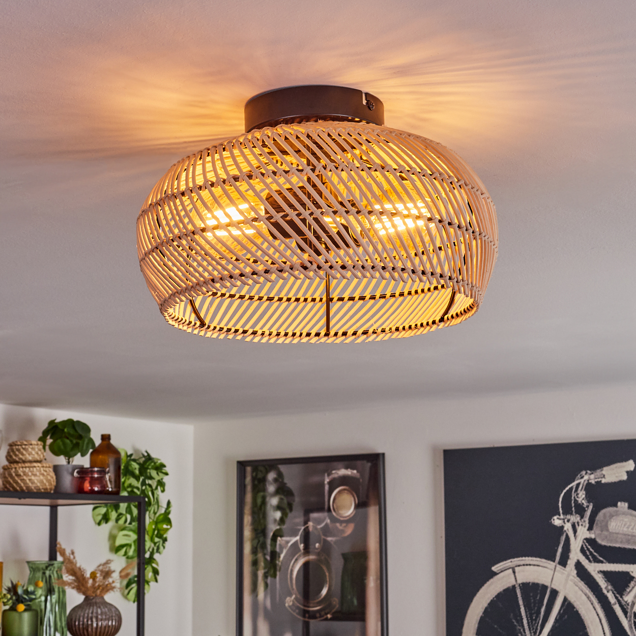 Eine Deckenlampe aus Rattan ist beleuchtet und wirft einen warmen Schein. Darunter sind ein Regal mit Pflanzen und Dekoration sowie Bilder an der Wand zu sehen.