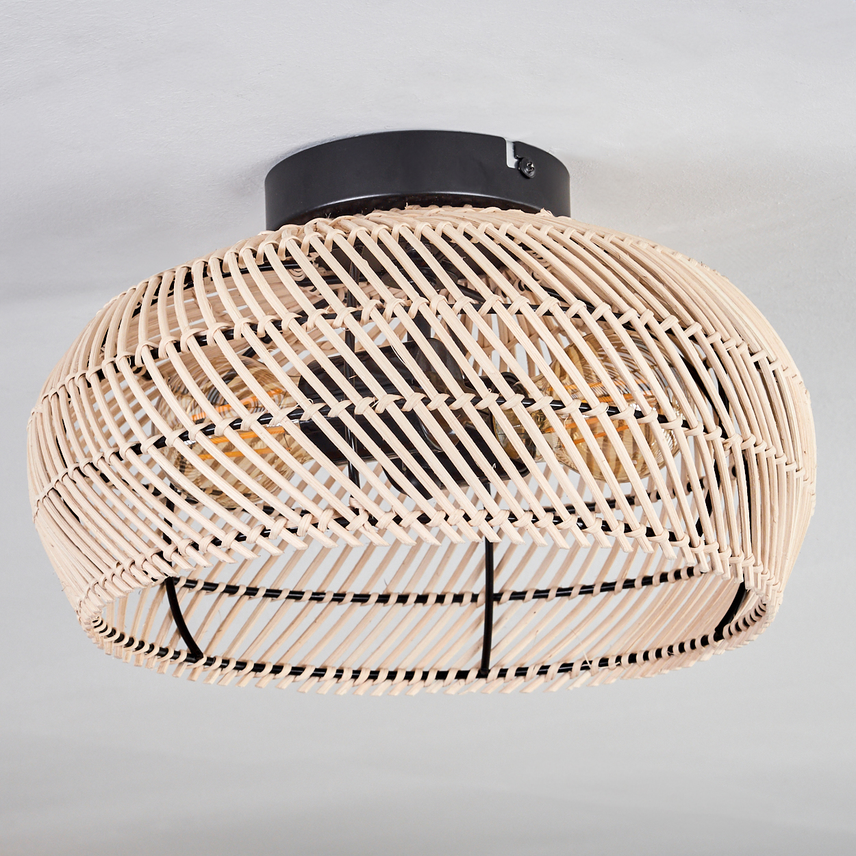 Eine Deckenleuchte aus geflochtenem Rattan mit schwarzem Sockel und Rahmen. Sie hat eine Glühbirne im Inneren und befindet sich vor weißem Hintergrund.