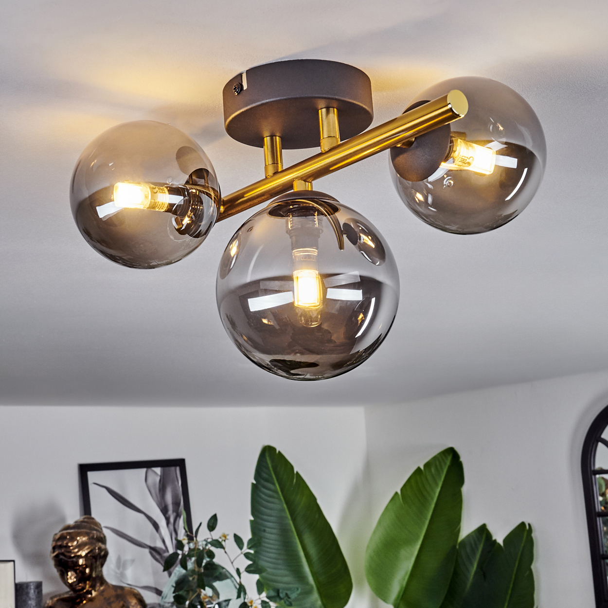 Eine moderne Deckenlampe mit drei rauchigen Glaskugeln, goldenen Armen und hellen Lichtern. Darunter grüne Blätter und Dekoration.