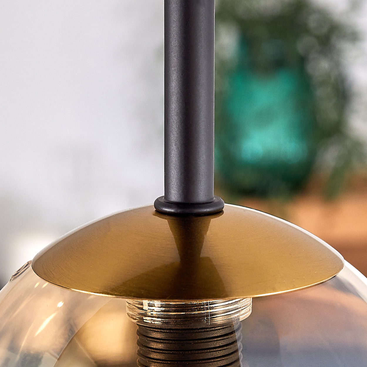 Nahaufnahme einer Pendelleuchte. Sie hat einen schwarzen Metallschaft, einen goldfarbenen Schirm und eine transparente Glühbirne. Der Hintergrund ist unscharf.