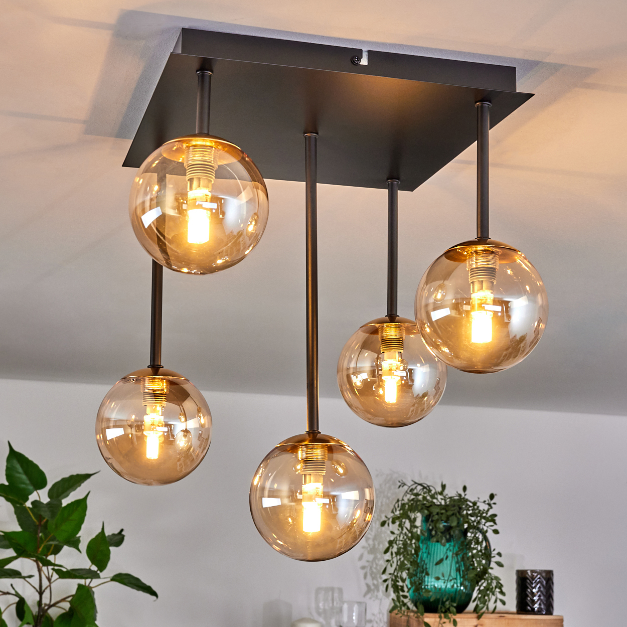 Eine moderne Deckenlampe mit fünf bernsteinfarbenen Glaskugeln. Die Lampe hängt an einer schwarzen quadratischen Basis und beleuchtet den Raum.