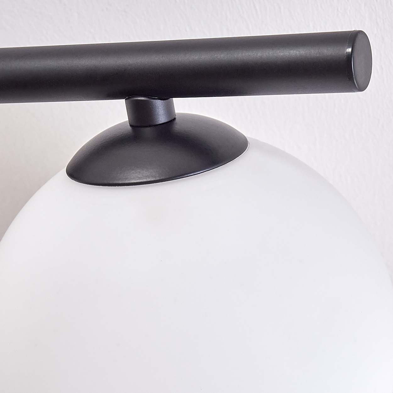 Nahaufnahme einer Lampe. Ein weißer, abgerundeter Glasschirm befindet sich unter einer schwarzen Metallhalterung und -stange an einer weißen Wand.