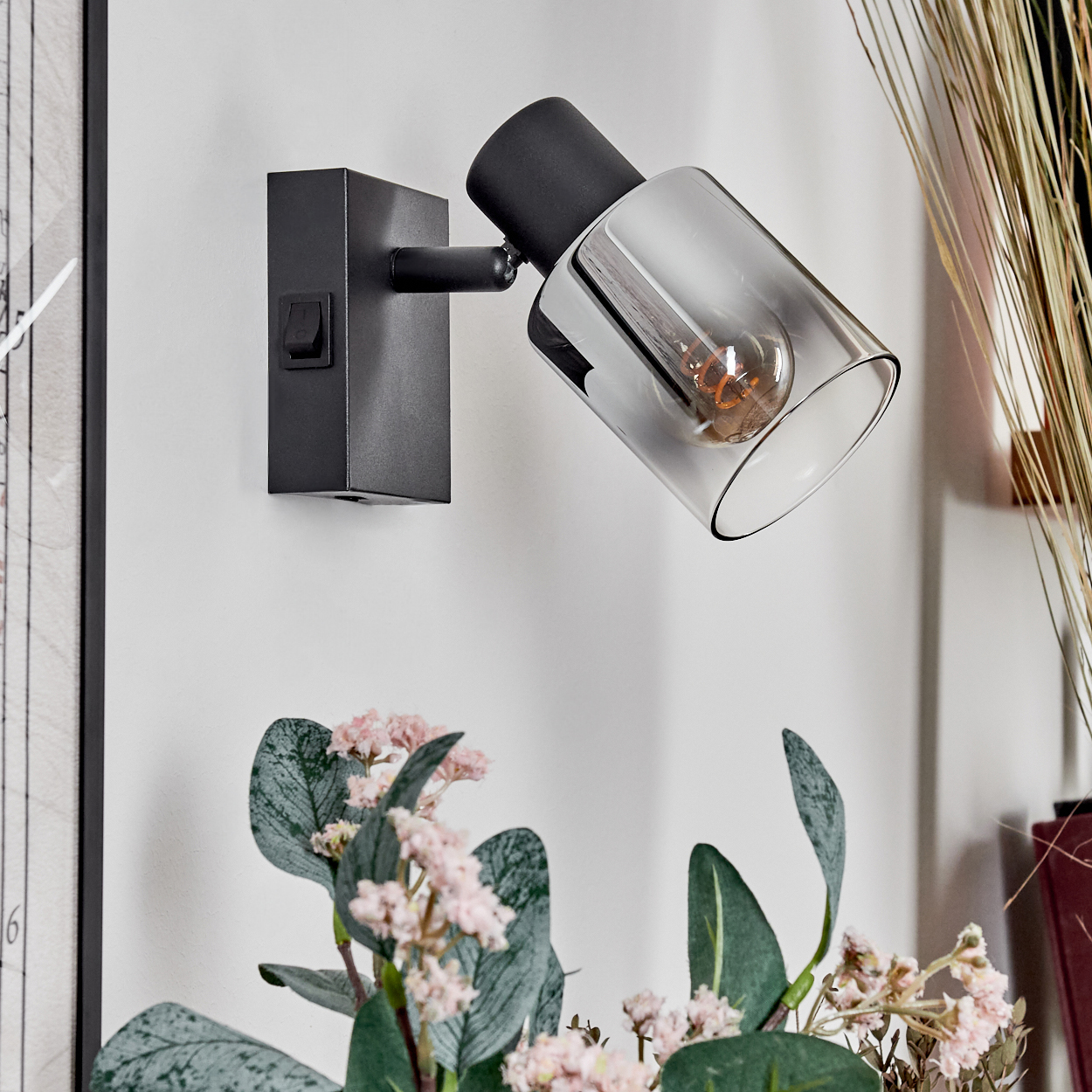 Eine schwarze Wandleuchte mit klarem Glasschirm ist an einer weißen Wand montiert. Rosa Blumen sind im Vordergrund.