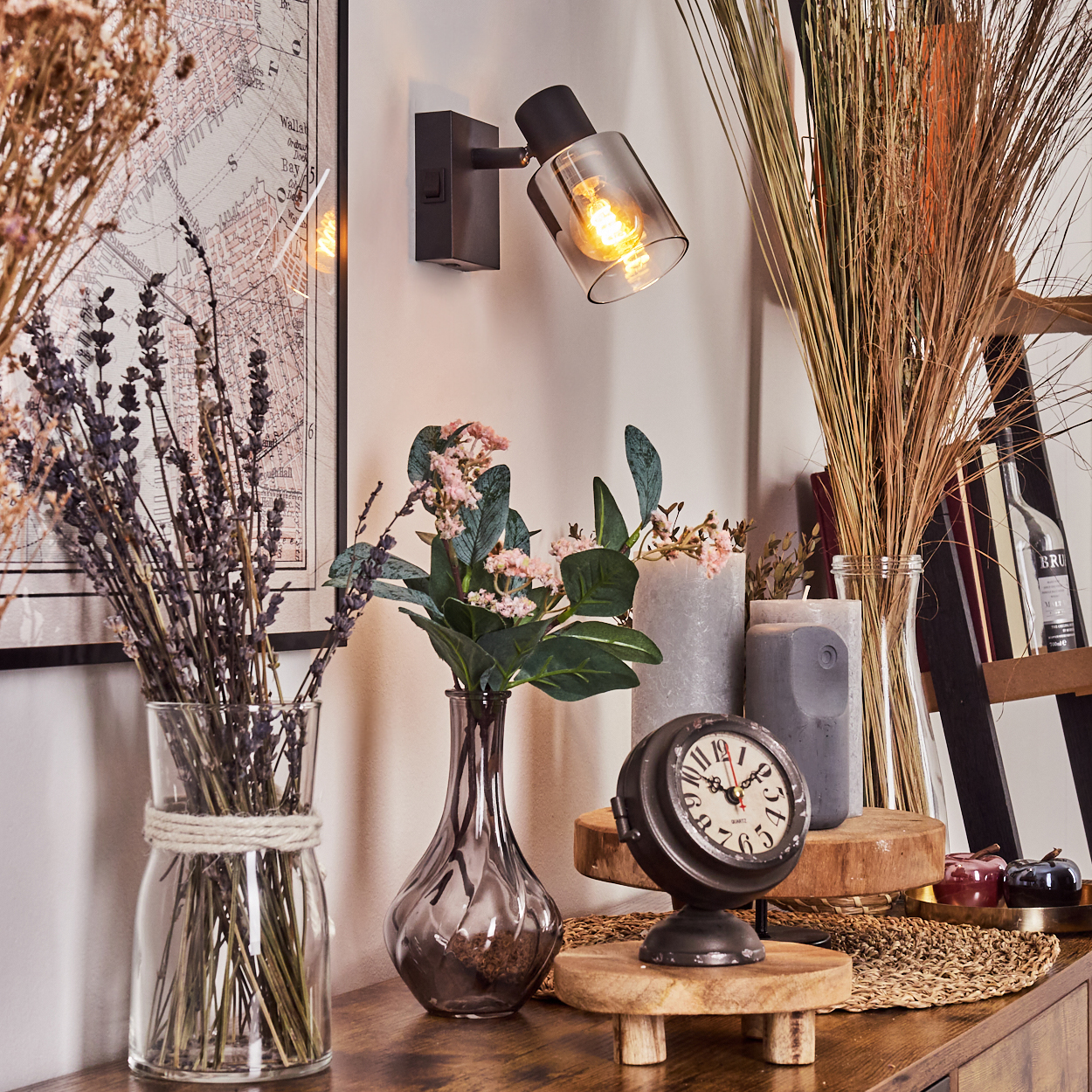 Ein Stillleben mit Trockenblumen, einer Uhr, Kerzen und einer Wandlampe auf einer Holzoberfläche. Im Hintergrund ist eine Karte.