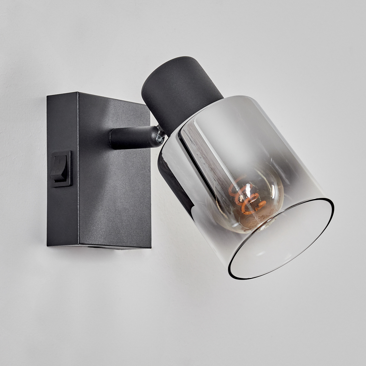 Eine schwarze Wandlampe mit zylindrischem Schirm. Sie hat einen Schalter und eine sichtbare Glühbirne. Der Hintergrund ist eine weiße Wand.