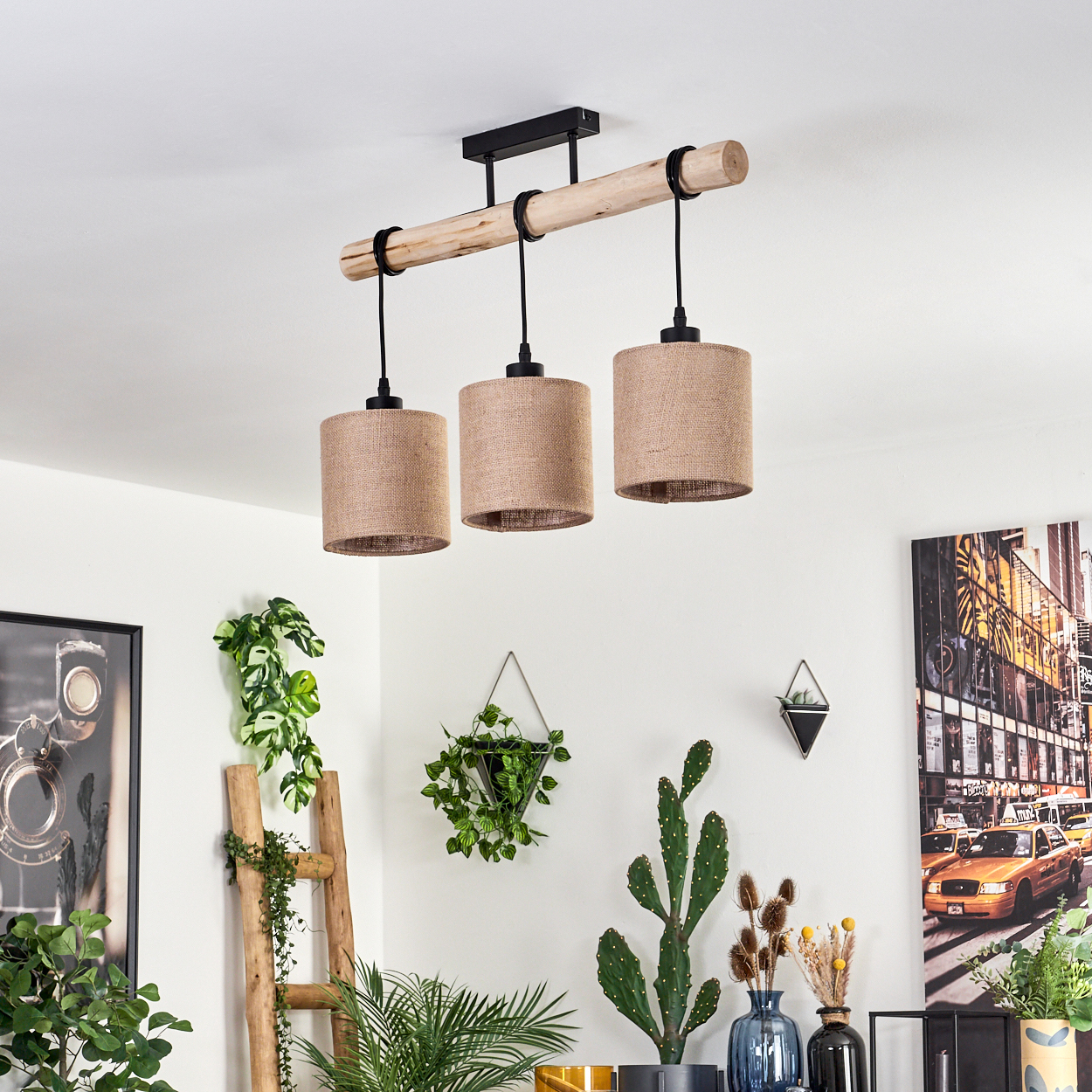 Eine moderne Deckenlampe mit drei Schirmen. Pflanzen und Bilder schmücken die weiße Wand. Eine Leiter und ein Kaktus befinden sich im Raum.