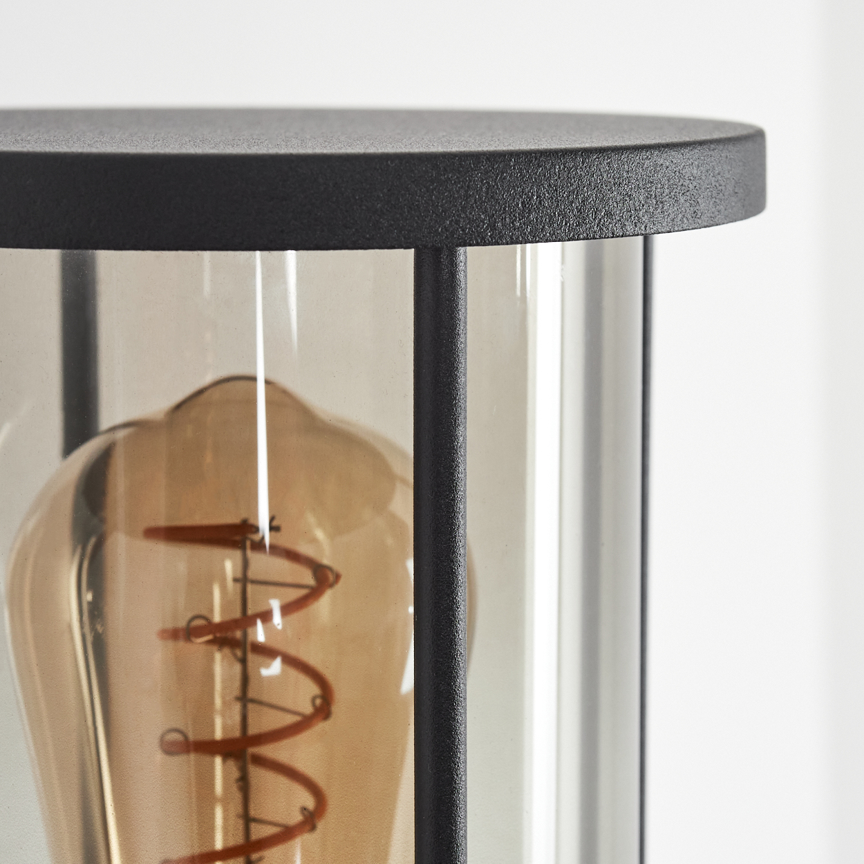 Nahaufnahme einer schwarzen Außenlampe. Sie hat eine Glasabdeckung mit einer warm getönten Glühbirne. Der Hintergrund ist weiß.