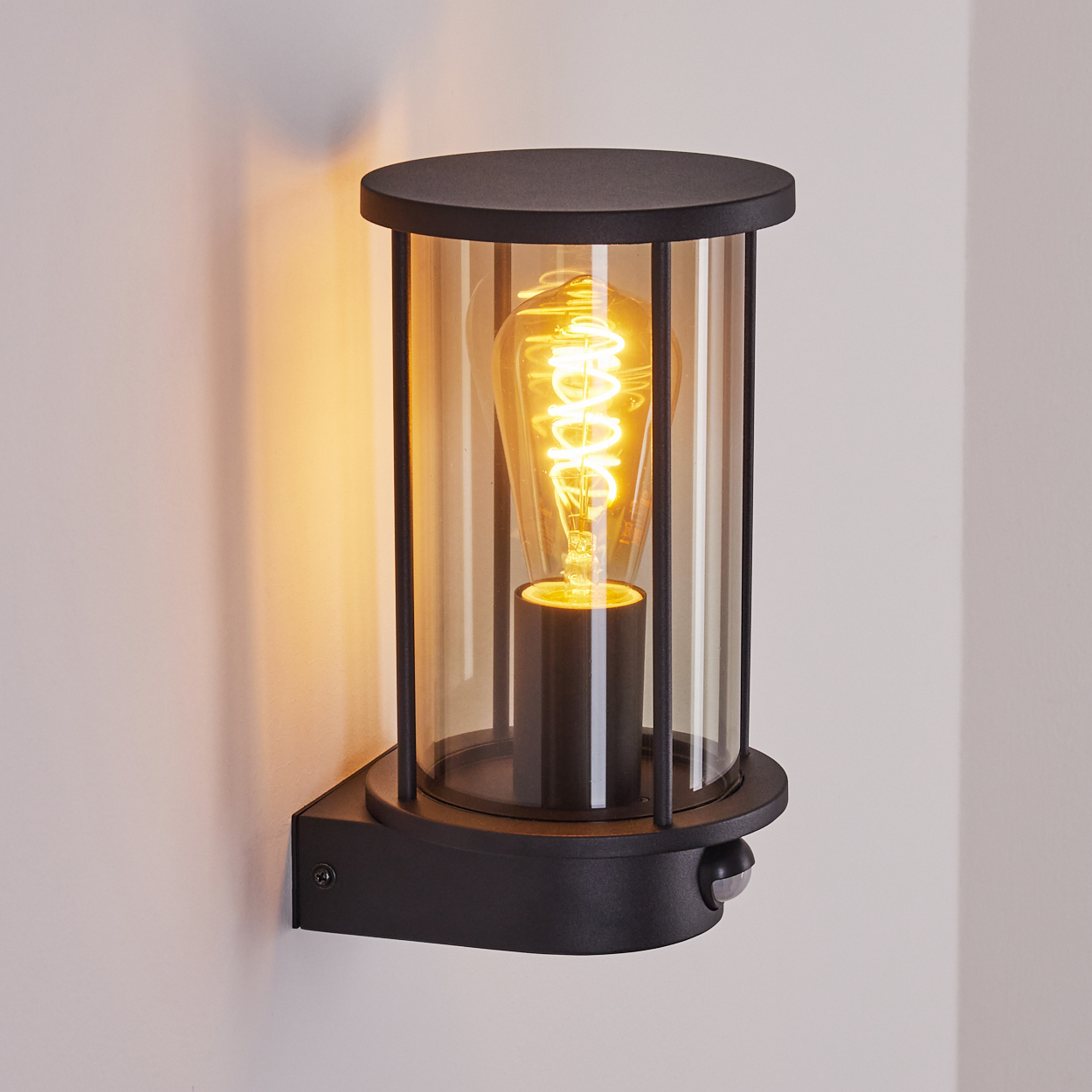 Schwarze Wandlampe mit einer leuchtenden Glühbirne in einem Glaszylinder. Die Lampe ist an einer weißen Wand mit Sensor befestigt.