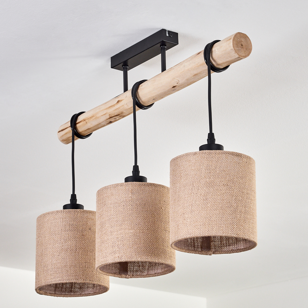 Eine Deckenleuchte mit drei Lampen mit Juteschirmen, die von einem Holzbalken hängen, der an einer schwarzen Metallplatte befestigt ist.
