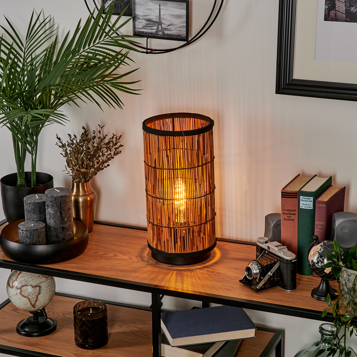 Eine Lampe mit Bambusschirm, beleuchtet. Bücher, Kamera, Globus und Pflanzen auf einem Holzregal. Kerzen und Rahmen an der Wand.