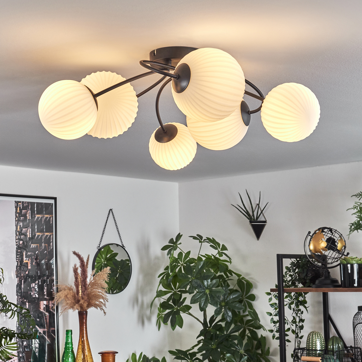 Deckenleuchte mit kugelförmigen Schirmen und dunklem Metallrahmen. Pflanzen und Dekorationen sind im Hintergrund zu sehen.