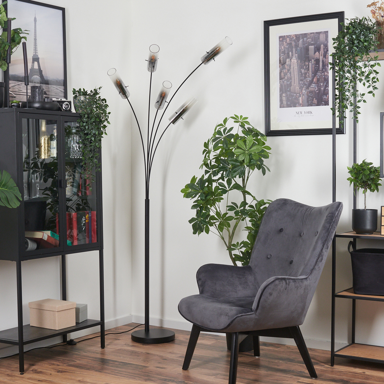 Ein Wohnzimmer mit grauem Sessel, schwarzer Stehlampe, Schrank und gerahmten Bildern. Pflanzen sind ebenfalls zu sehen.