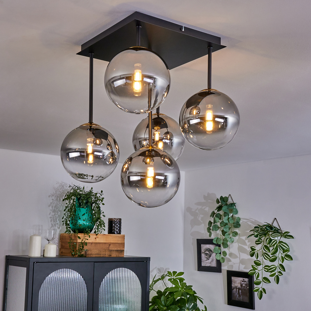 Eine moderne Deckenlampe mit fünf Glaskugeln hängt von der Decke. Darunter steht ein Schrank mit Pflanzen und Dekoration an einer weißen Wand.