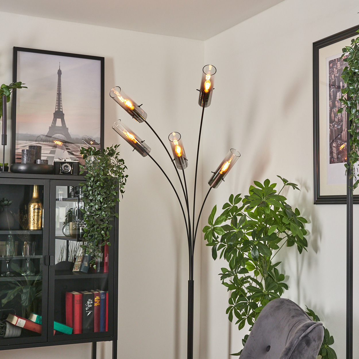 Ein modernes Wohnzimmer mit Stehlampe, Schrank und gerahmten Bildern. Pflanzen und ein grauer Sessel sind ebenfalls zu sehen.