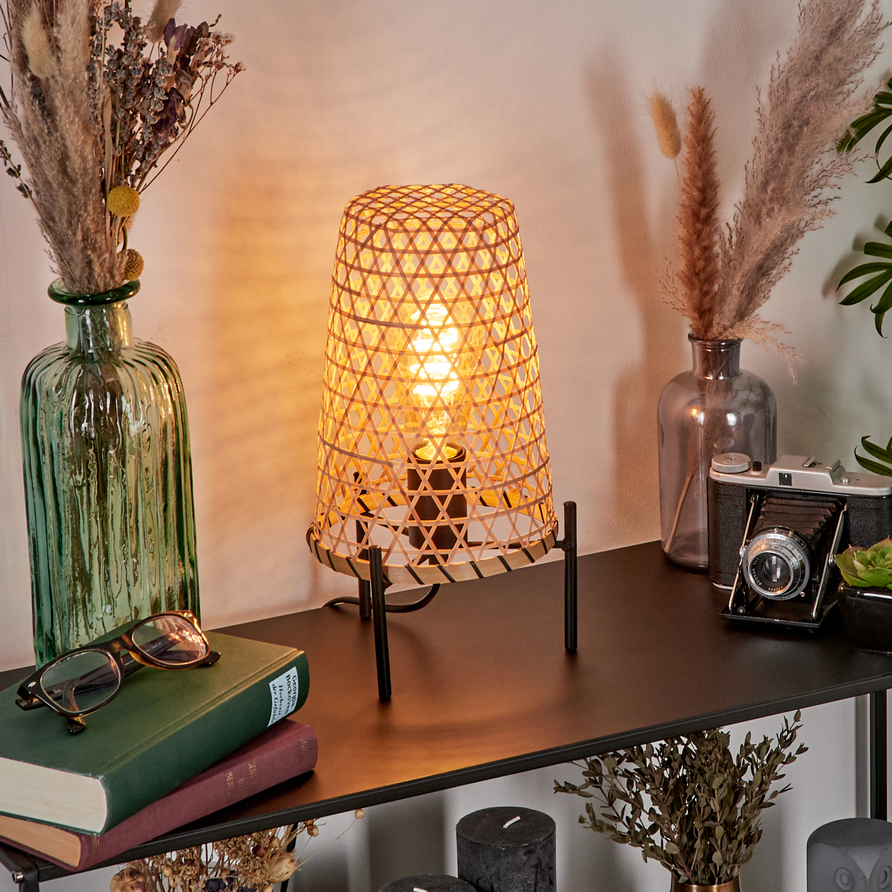 Gewebte Lampe auf schwarzem Regal mit Büchern, Gläsern, Vasen mit Trockenblumen, einer Kamera und Kerzen.