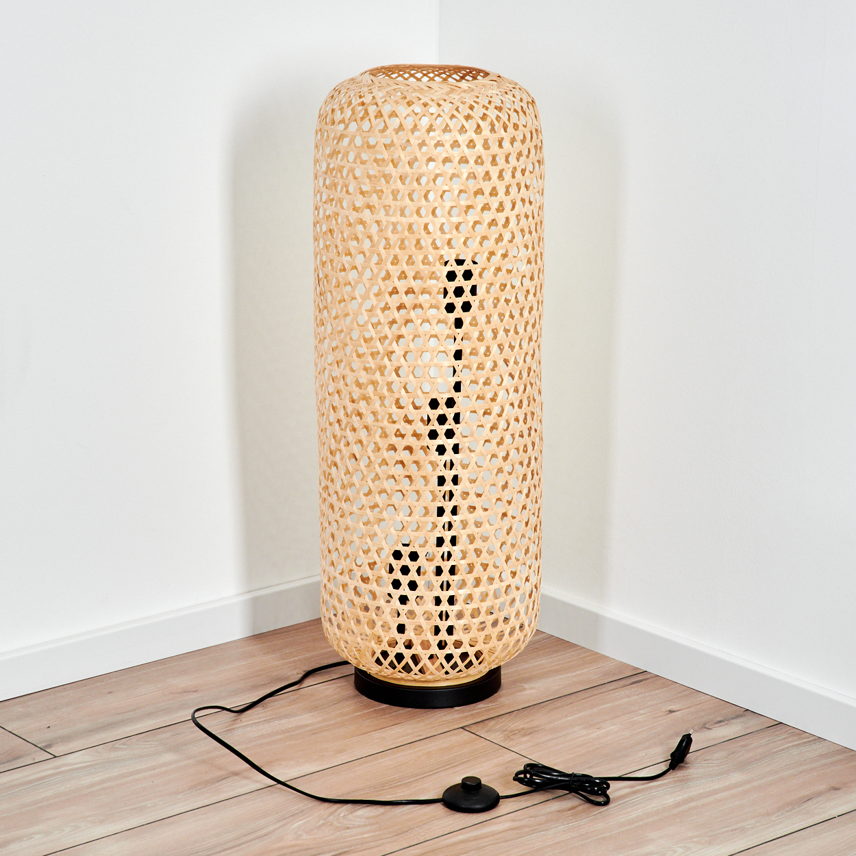 Eine gewebte Bambus-Stehlampe steht in einer Ecke. Sie hat einen schwarzen Sockel und ein Netzkabel. Der Boden ist aus Holz.