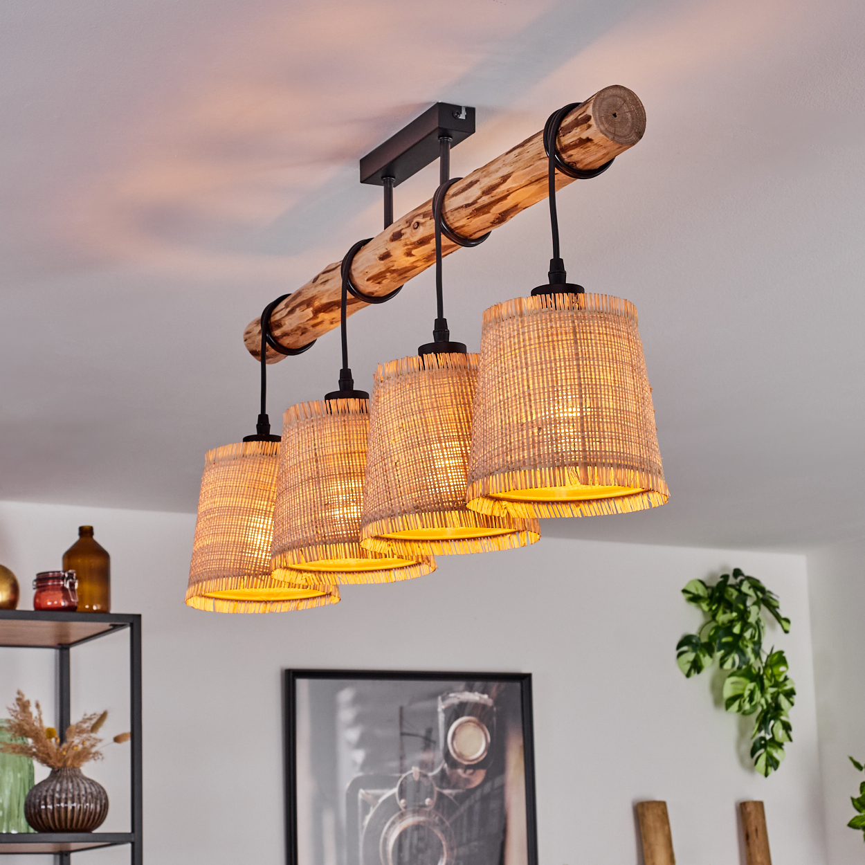 Ein Holzbalken mit vier gewebten Lampen hängt von der Decke. Die Lampenschirme sind beige und das Licht ist warm. Dekoartikel sind im Hintergrund.