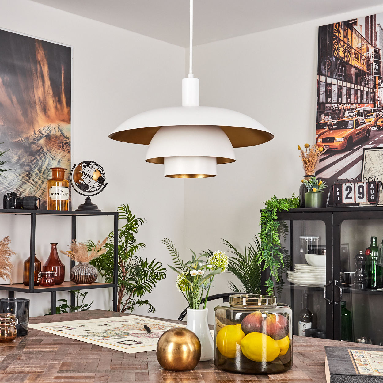 Ein Esszimmer mit einer weißen und goldenen Lampe. Tisch mit Früchten, Globus und Pflanzen. Kunstwerk an der Wand. Schwarzes Regal mit Dekor.