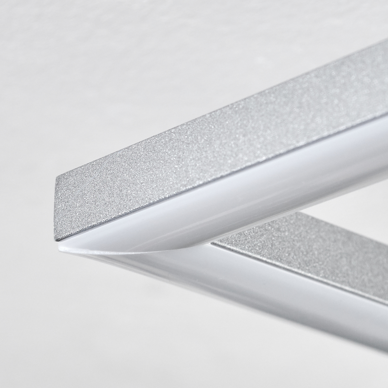 Nahaufnahme einer metallischen, rechteckigen Leuchte. Die Kanten sind strukturiert silberfarben, mit einer weißen Innenfläche. Der Hintergrund ist schlicht weiß.