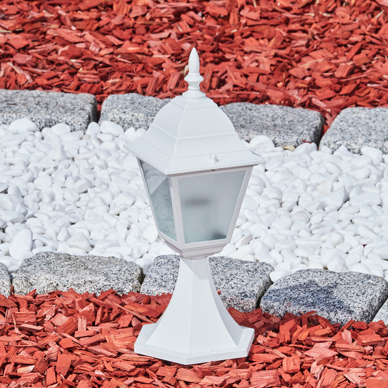 Eine weiße Außenlampe steht auf roter Mulche, umgeben von weißen Steinen und grauen Felsen. Der Hintergrund ist rote Mulche.