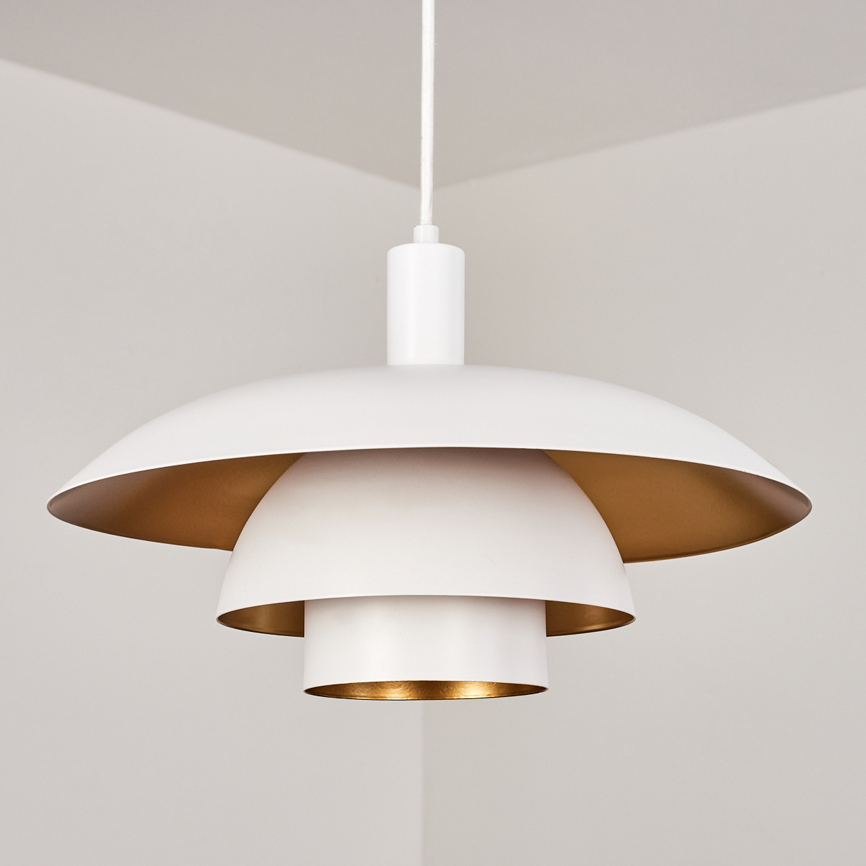 Eine weiße Hängeleuchte mit mehrschichtigem Design und goldenen Innenräumen. Sie hängt in einer Ecke von der Decke.