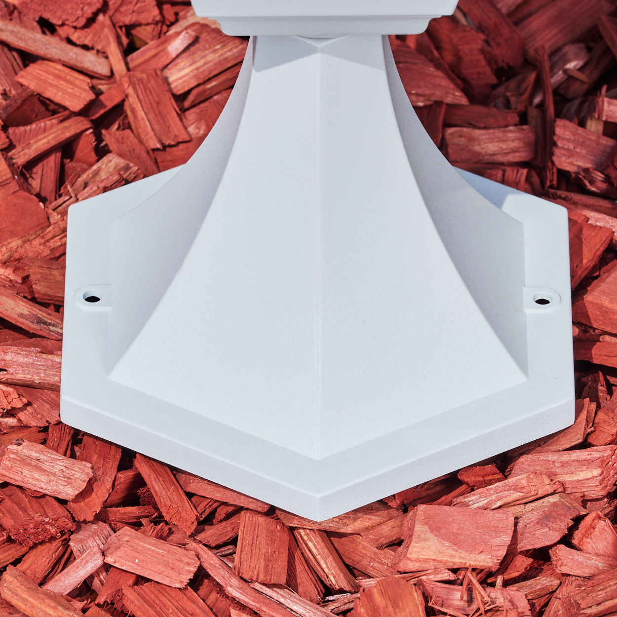 Ein weißer, geometrischer Sockel steht auf roter Mulch. Die Basis hat eine sechseckige Form und zwei Löcher. Der Hintergrund zeigt Holzspäne.