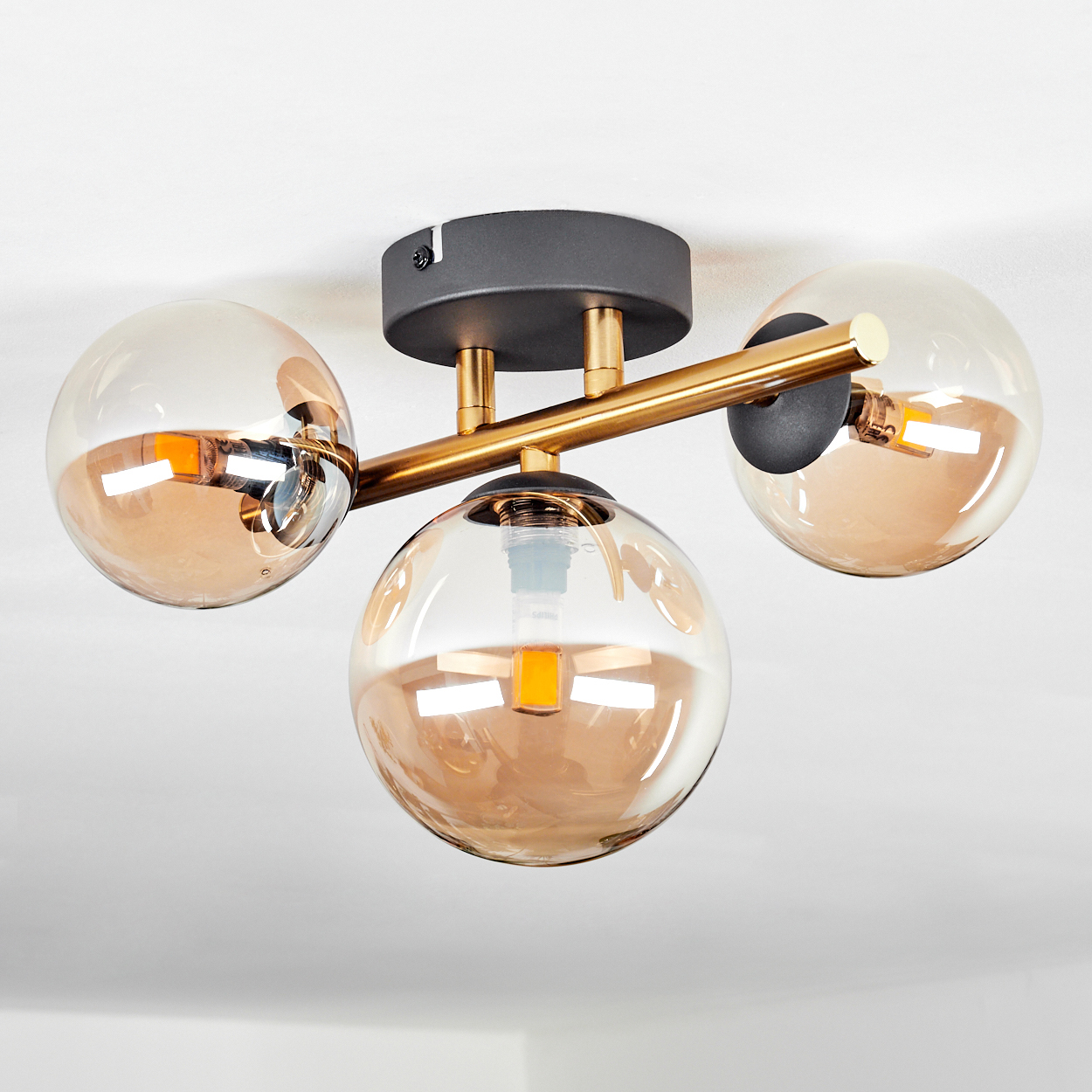 Eine Deckenlampe mit drei kugelförmigen Glasschirmen. Goldakzente und ein schwarzer Sockel. Die Schirme sind hell bernsteinfarben.
