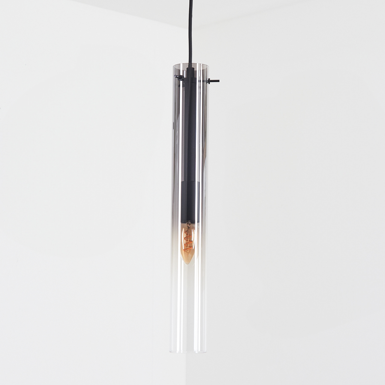 Eine Hängelampe mit zylindrischem Glasschirm hängt an einem schwarzen Kabel vor weißem Hintergrund. Die Glühbirne ist sichtbar.