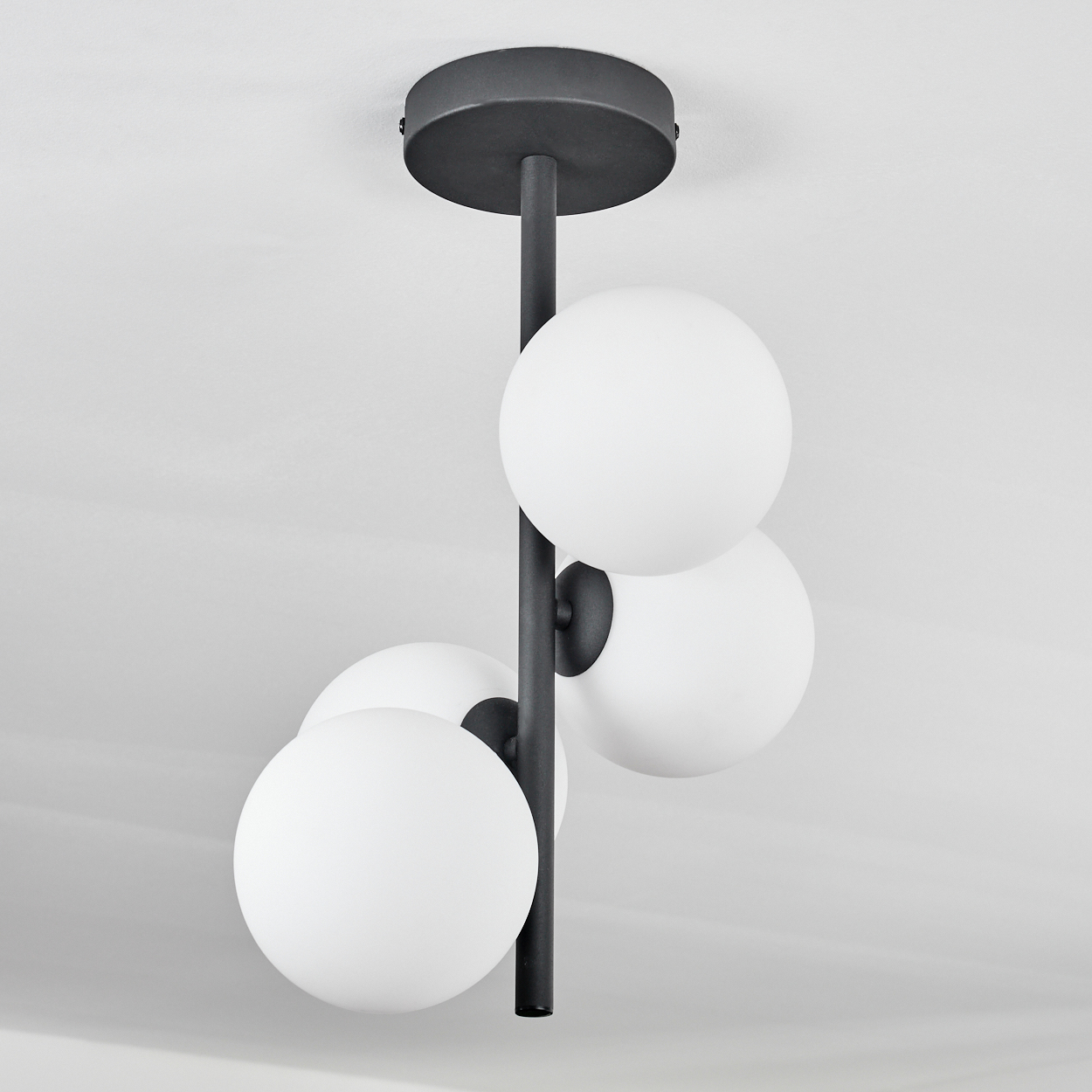 Eine moderne Deckenlampe mit vier weißen, kugelförmigen Glühbirnen, die an einer schwarzen Stange befestigt sind. Sie ist an einer weißen Decke montiert.