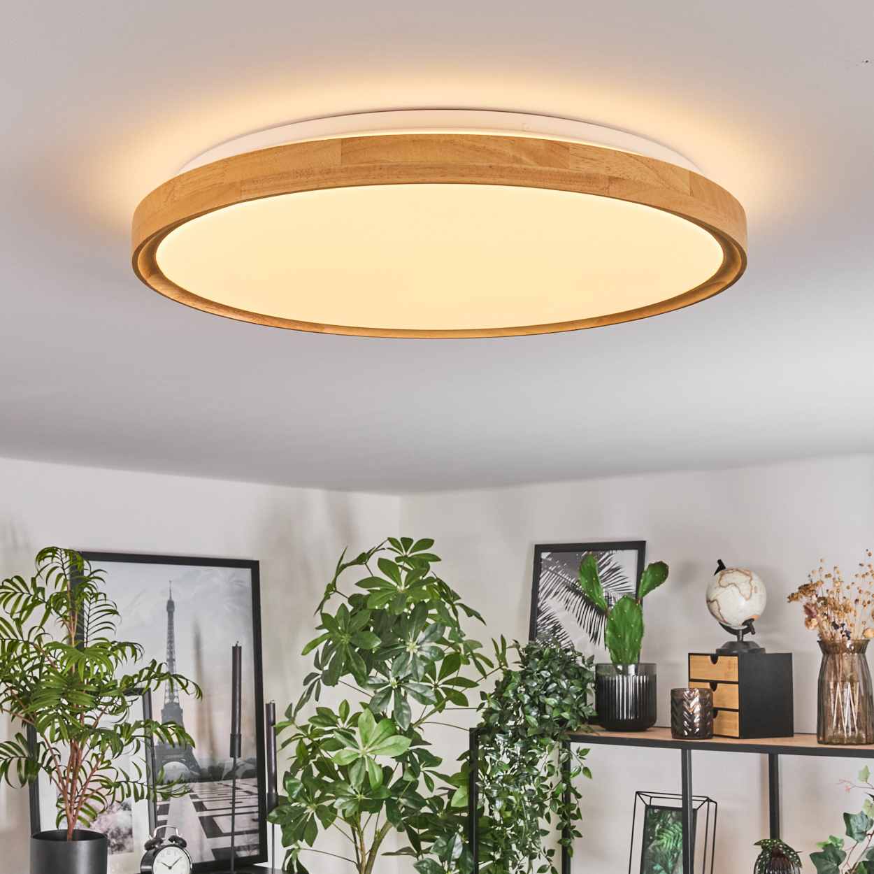 Eine runde Deckenlampe aus Holz beleuchtet einen Raum mit Pflanzen, Bildern und Dekoration. Die Lampe hat einen weißen Innenkreis.