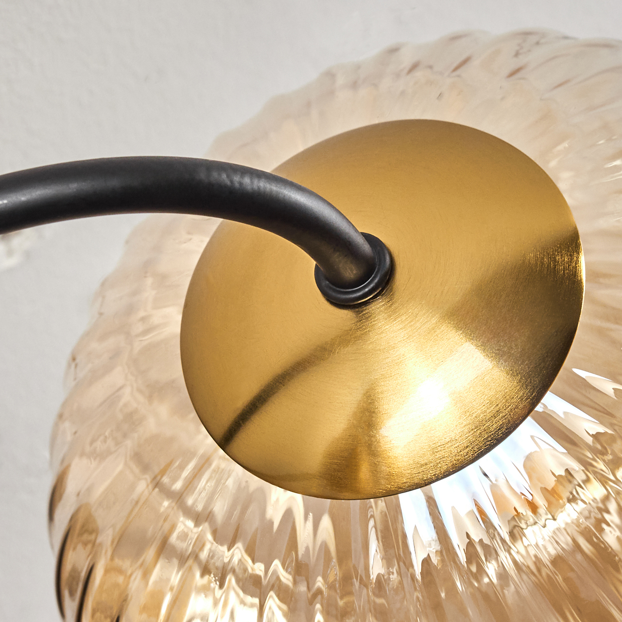 Nahaufnahme einer Leuchte. Sie hat einen goldfarbenen Sockel, einen gebogenen schwarzen Arm und einen strukturierten, bernsteinfarbenen Glasschirm.