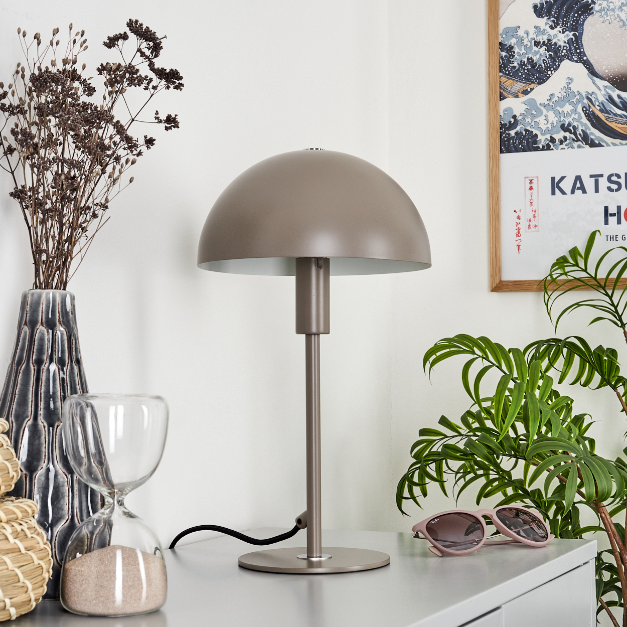 Ein Schreibtisch mit Lampe, Vase mit trockenen Pflanzen, Sanduhr, Sonnenbrille und Gemälde. Grüne Pflanze rechts.