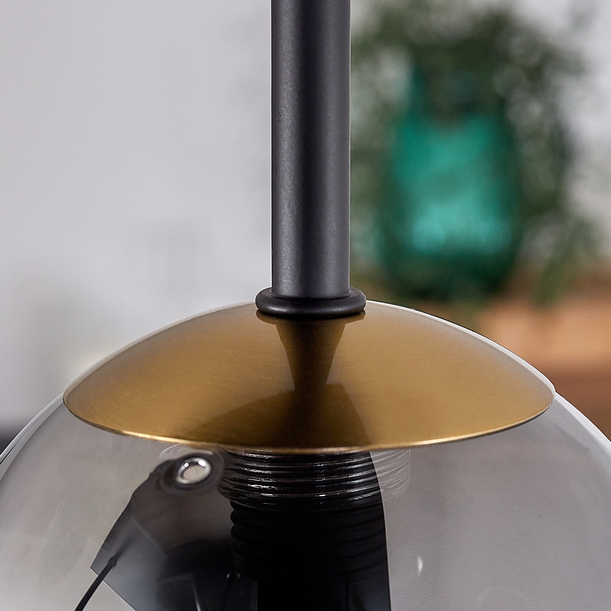 Nahaufnahme einer modernen Lampe. Sie hat einen schwarzen Stiel, einen goldenen Schirm und eine graue Glühbirne. Im Hintergrund ist eine grüne Vase unscharf.