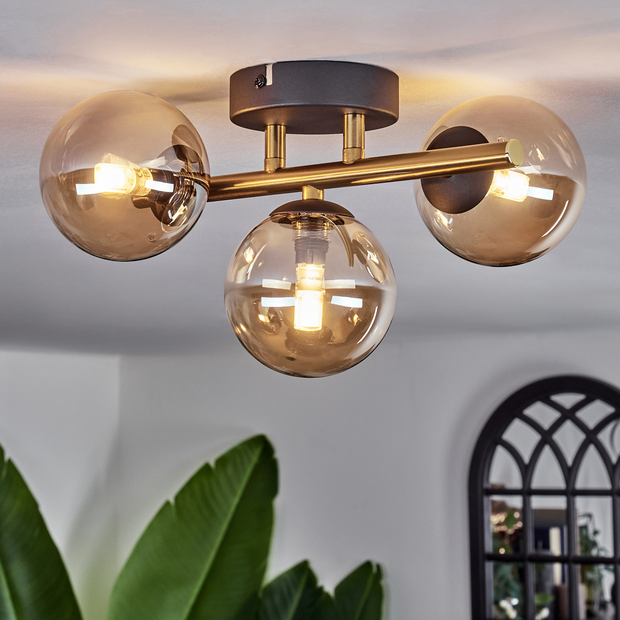 Deckenlampe mit drei Glaskugeln. Jede Kugel enthält eine brennende Glühbirne. Der Rahmen ist messingfarben, mit dunklergrauem Sockel. Pflanze sichtbar.