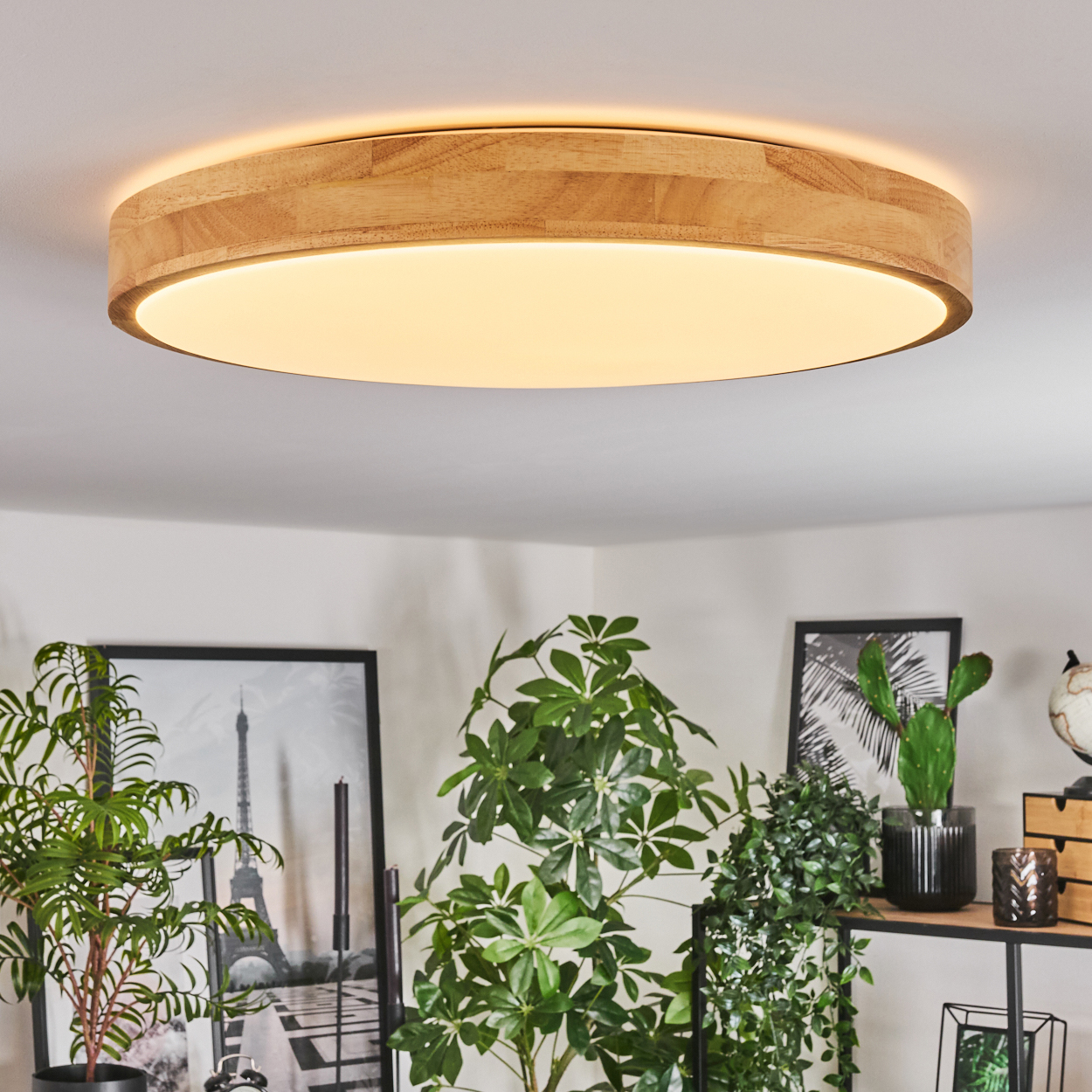 Eine runde Deckenleuchte aus Holz beleuchtet einen Raum mit Pflanzen und gerahmten Bildern. Das Licht ist von einem Holzrahmen umgeben.