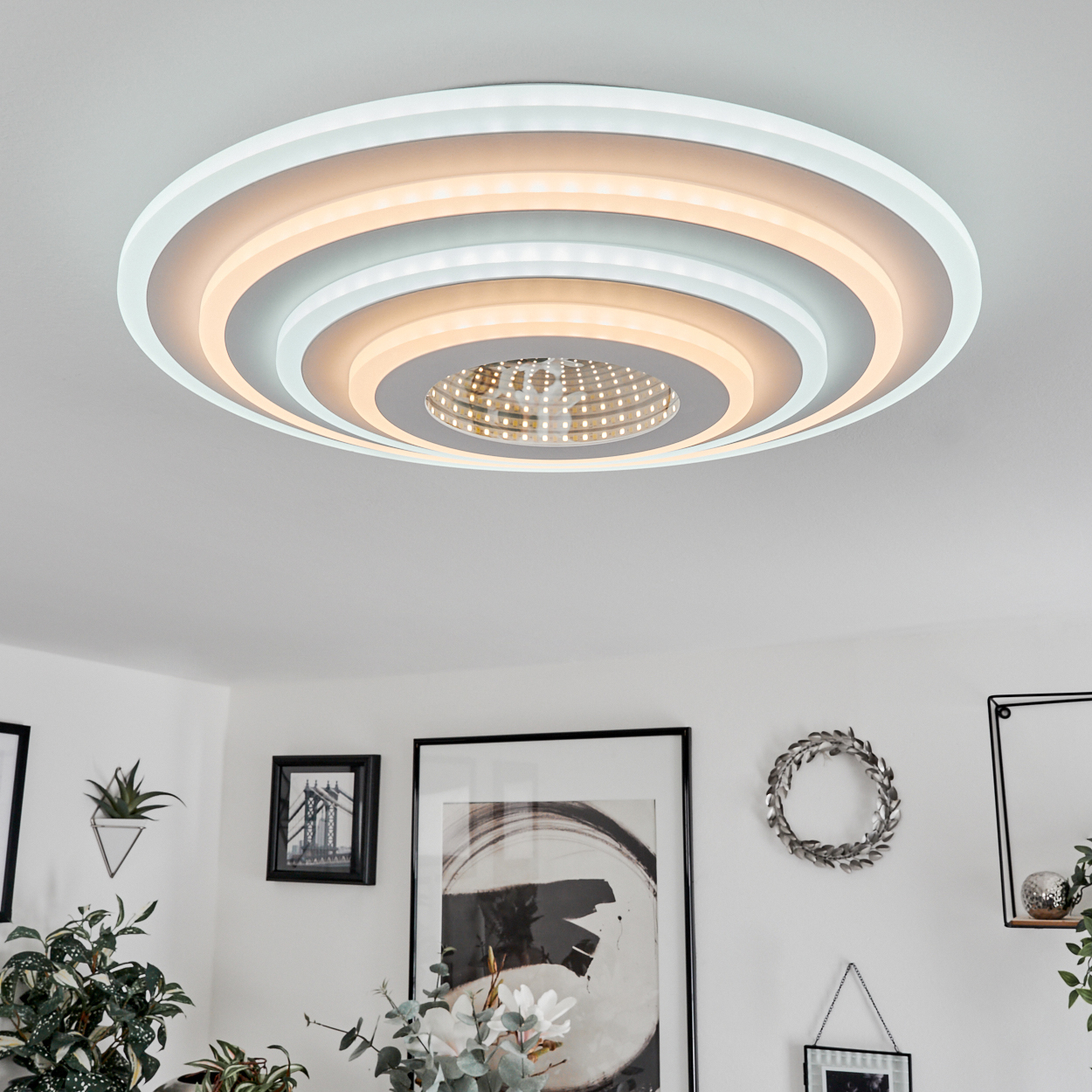 Eine moderne Deckenlampe mit konzentrischen Kreisen, die Licht emittieren. Darunter ist ein Raum mit Kunst und Pflanzen sichtbar.