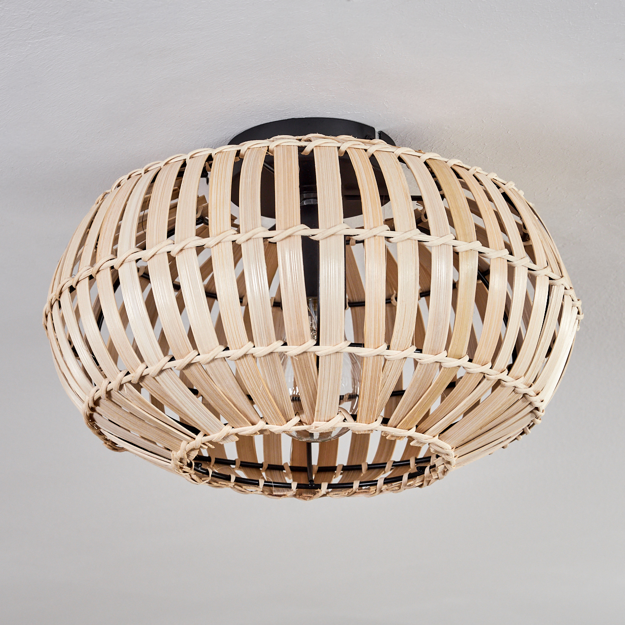 Ein gewebter Bambus-Lampenschirm ist an einer weißen Decke befestigt. Die Lampe ist kreisförmig mit schwarzem Sockel und Glühbirne.