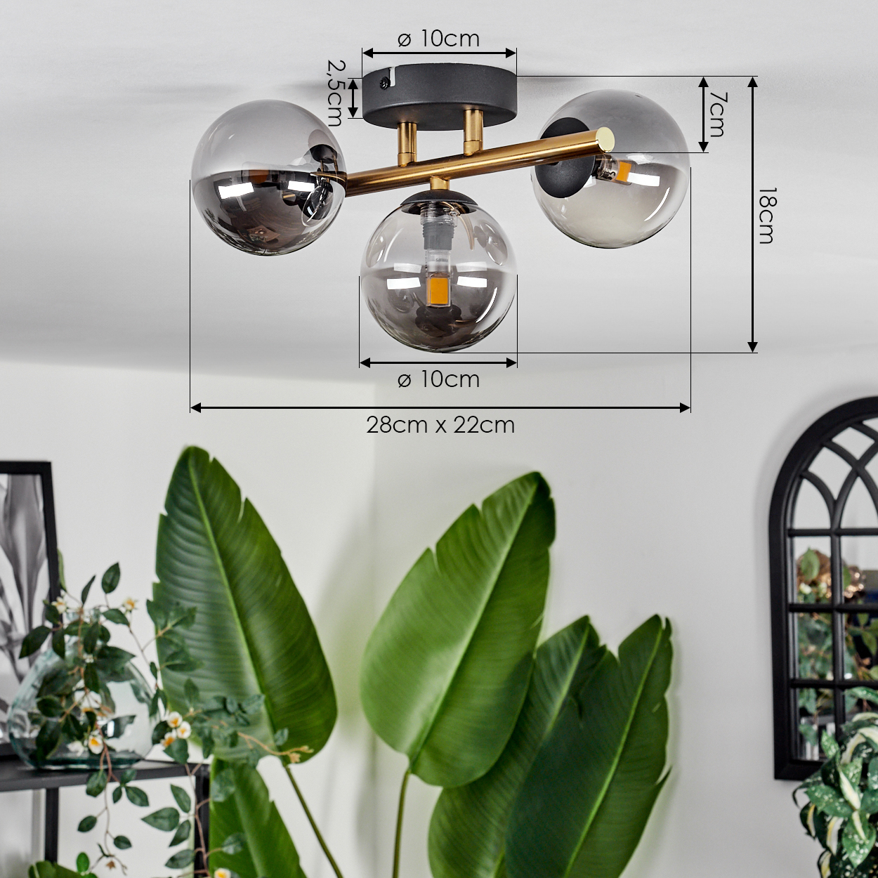 Eine moderne Deckenlampe mit drei Glaskugeln. Im Hintergrund sind große grüne Blätter und ein Fenster zu sehen.